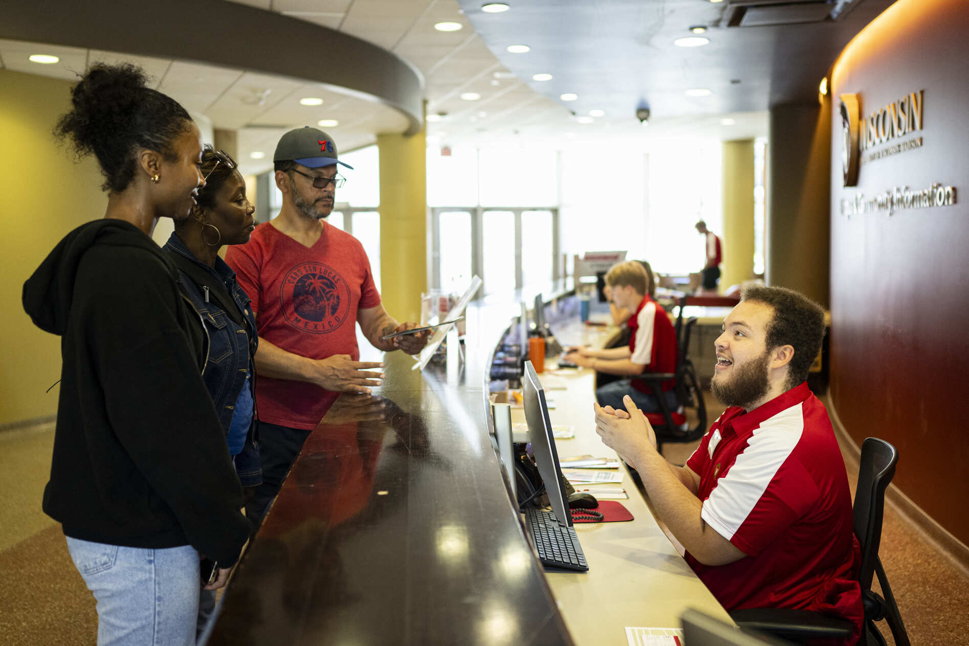Three visitors stand at a welcome desk as student staff members in red-and-white shirt assists them.