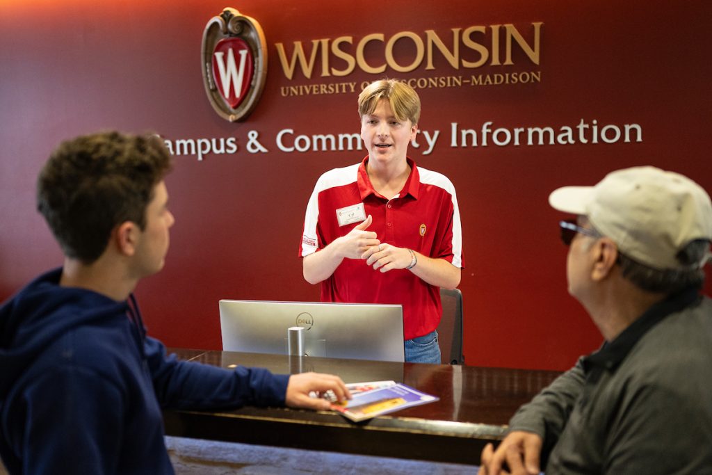 A student information guide speaks with two visitors across an information desk, with a red “Campus & Community Information” wall behind them.