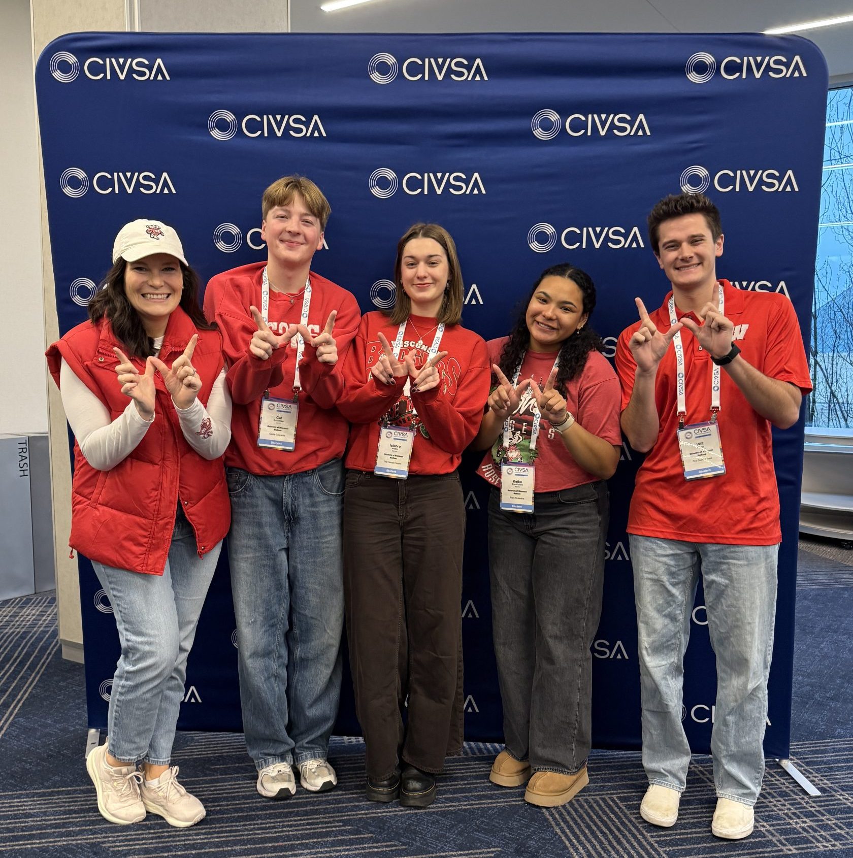 A small group of people in red shirts pose in front of a blue CIVSA backdrop, each holding up a “W” hand sign and wearing event lanyards.